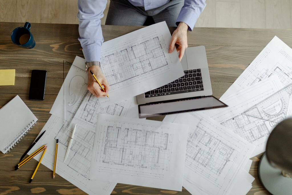 “Architects reviewing and marking up house plan blueprints on a desk with a laptop and drafting tools.”
