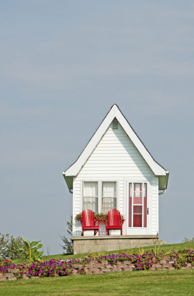 Small white tiny house with a front porch and red door on a grassy lot